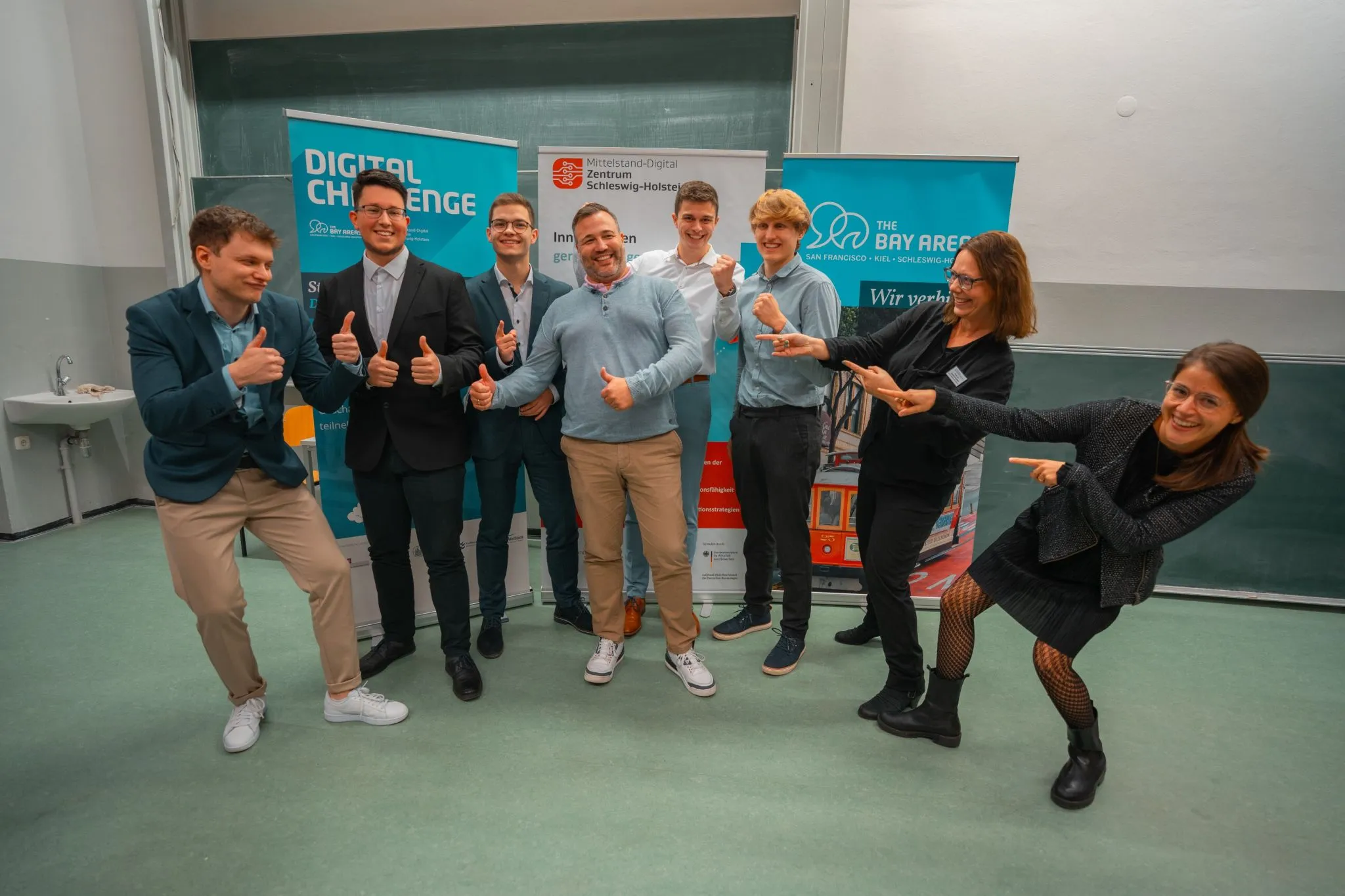 Sieben Männer und Frauen stehen gemeinsam mit IDALABS-Gründer Tobias Gürtler vor mehreren Bannern mit der Aufschrift “Digital Challenge”. Sie lachen und strecken ihre Daumen in die Höhe.