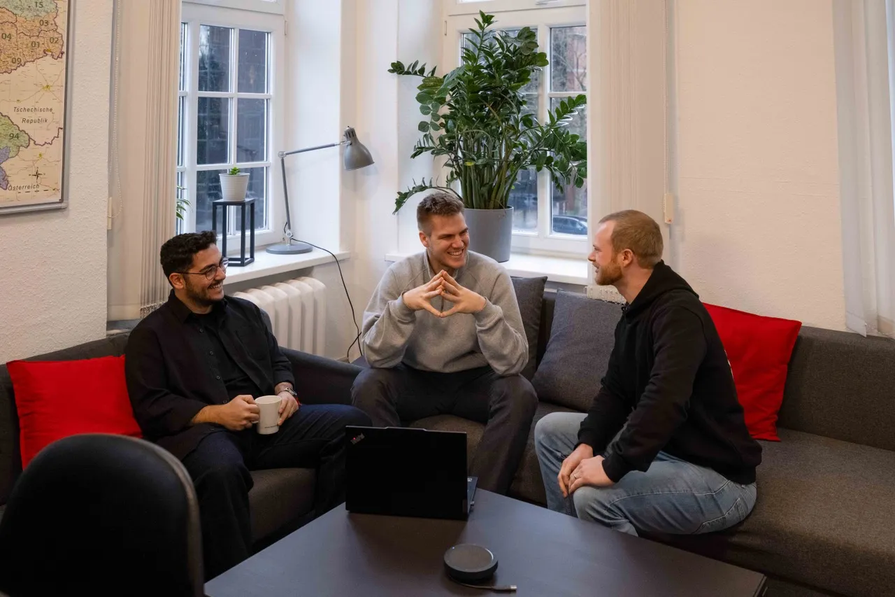 Drei junge Männer sitzen in einer Sofaecke im IDALABS-Büro und unterhalten sich fröhlich. Einer von ihnen hat einen Kaffeebecher in der Hand, zwischen ihnen steht ein schwarzer Laptop.