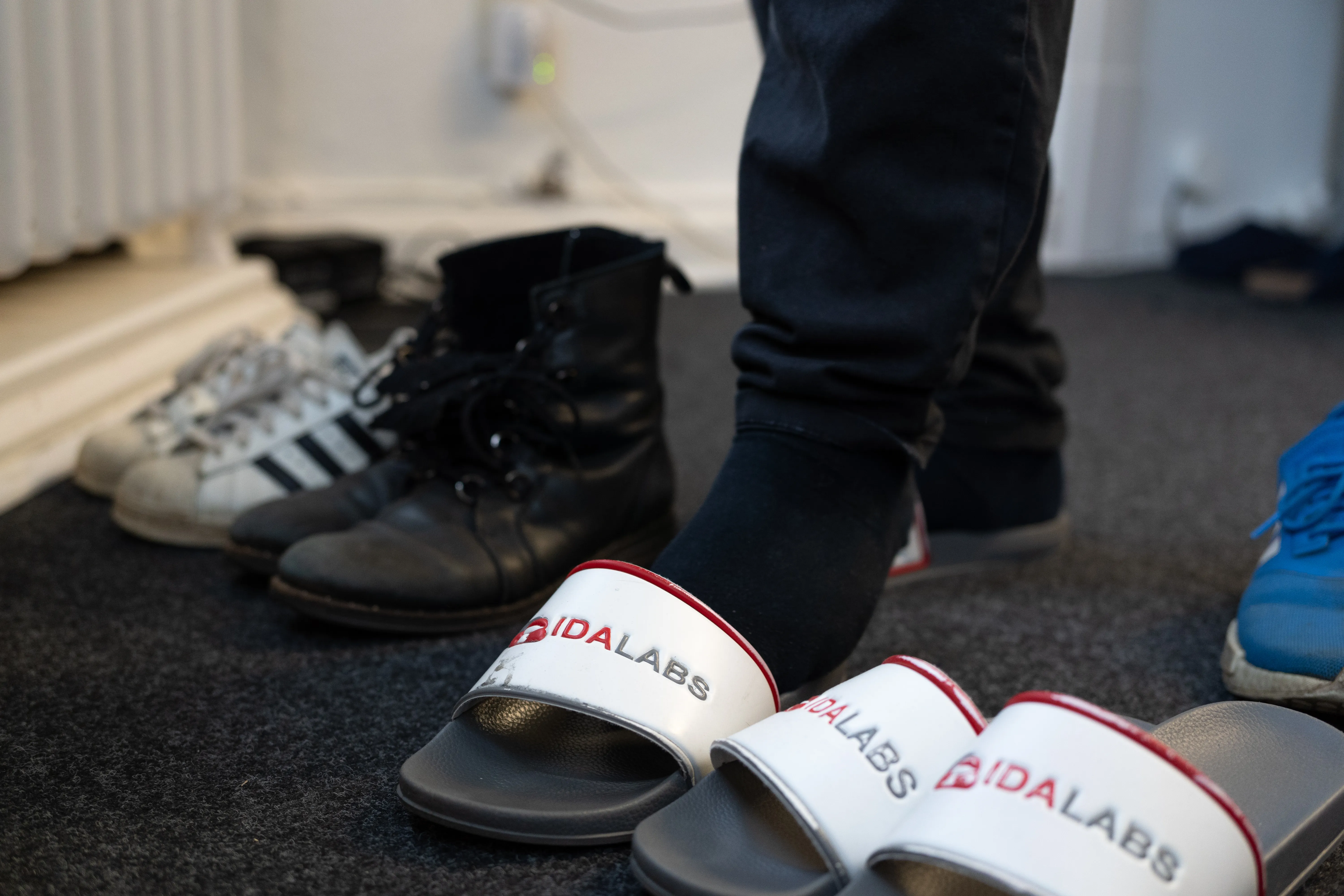 In einer Office-Garderobe stehen mehrere Paare Schuhe. Ein Mitarbeiter wechselt gerade von seinen Straßenschuhen in ein paar Badelatschen mit IDALABS-Aufdruck