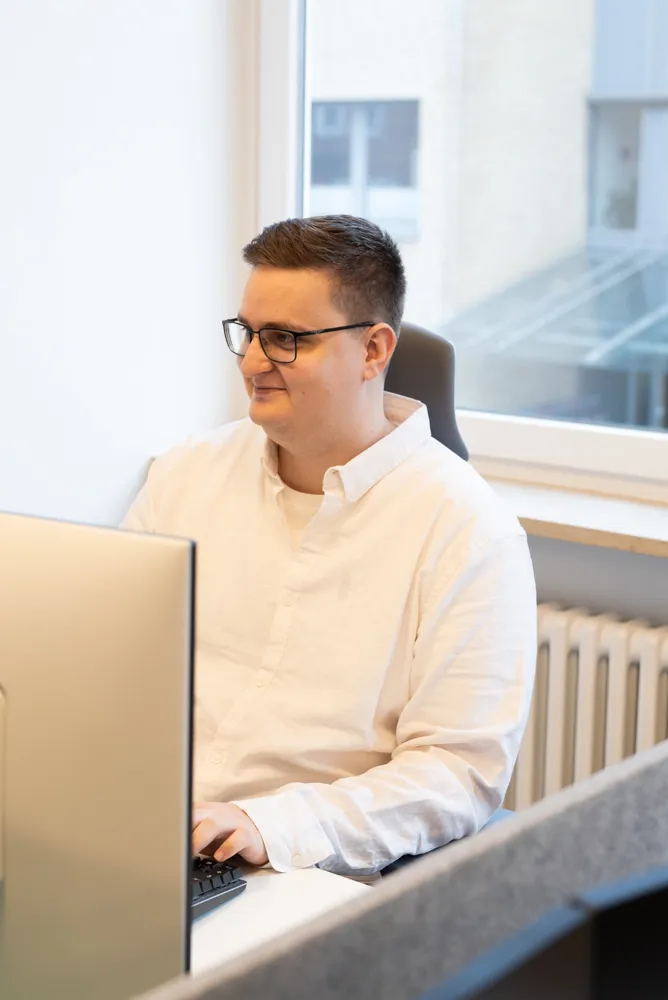 Ein junger Mann in weißem Hemd sitzt auf einem Schreibtischstuhl vor seinem Computer. Er schaut lächelnd auf den Bildschirm.