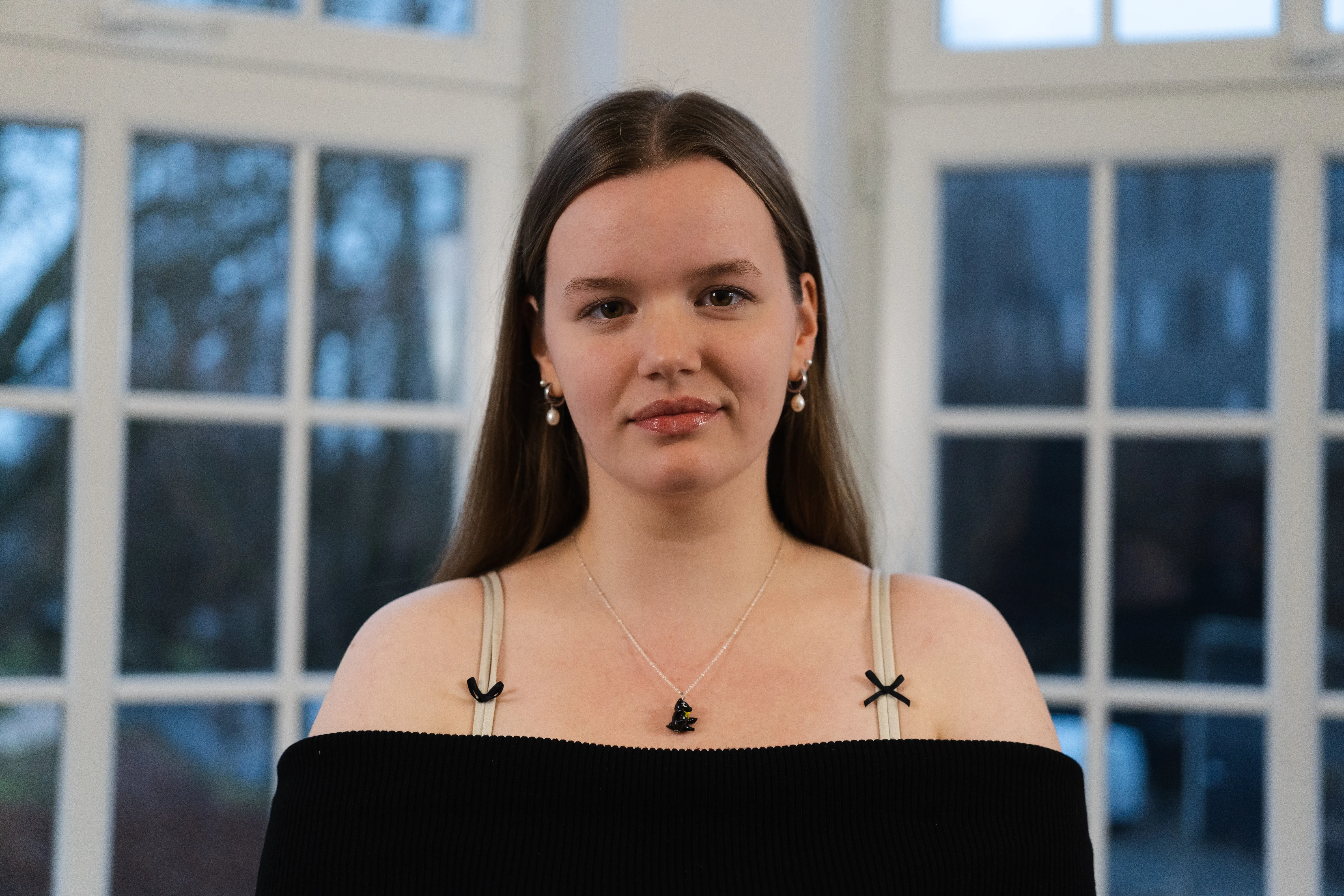 Electra Kausch schaut aufmerksam in die Kamera. Sie trägt lange braune Haare und einen schulterfreien, schwarzen Pullover