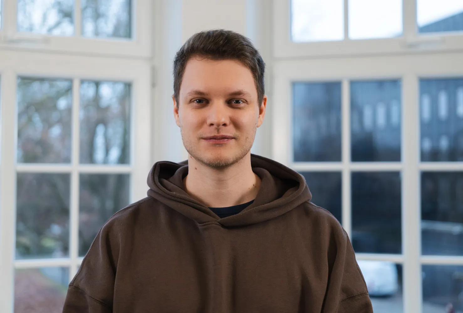 Adrian Frenz schaut freundlich in die Kamera. Er ist ein junger Mann mit braunen Haaren und blauen Augen. Er trägt einen braunen Kapuzenpullover.