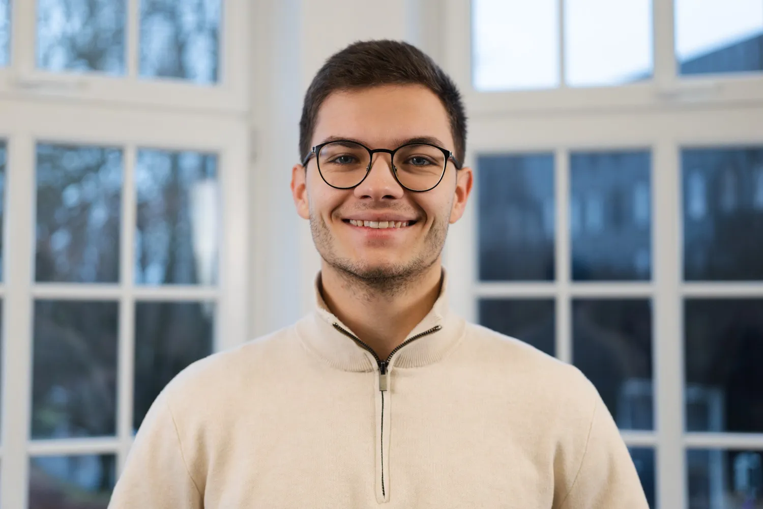 Lennart Leopold lacht freundlich in die Kamera. Er hat braune Haare und blaue Augen und trägt eine schwarze Brille sowie einen weißen Rollkragenpullover.