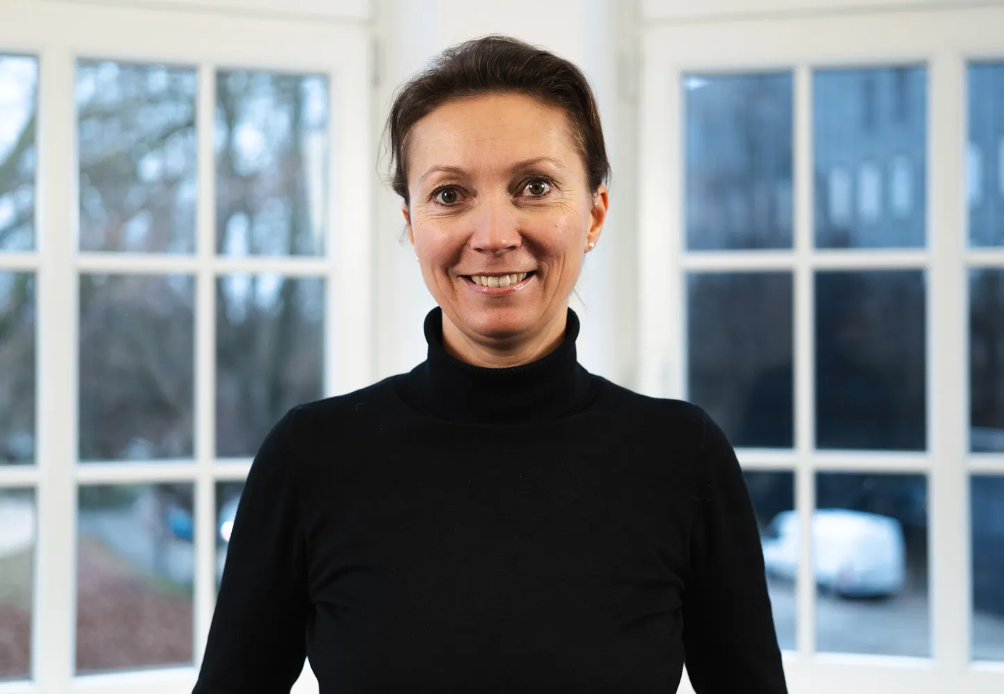 Sarah Gürtler steht vor einer Fensterfront und lächelt in die Kamera. Sie hat ihre braunen Haare zu einem Pferdeschwanz zusammengebunden und trägt einen schwarzen Rollkragenpullover.