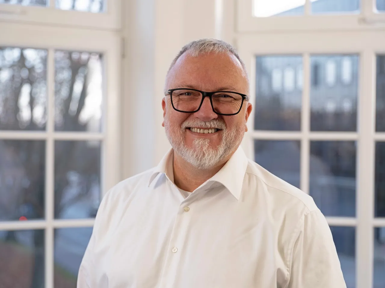 Ein Mann mit weißen Haaren und Bart steht vor den Fenstern eines Bürogebäudes und lacht in die Kamera. Er trägt eine Brille und ein weißes Hemd.