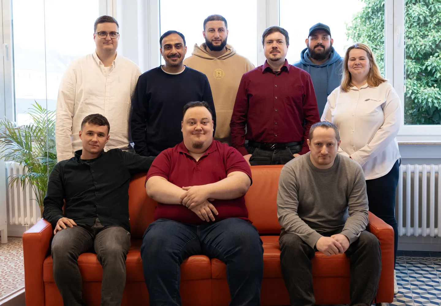 Drei junge Männer sitzen auf einem roten Sofa vor einer Fensterfront und schauen freundlich in die Kamera. Dahinter stehen fünf weitere junge Männer und eine junge Frau. Sie tragen bunte, legere Office-Kleidung.