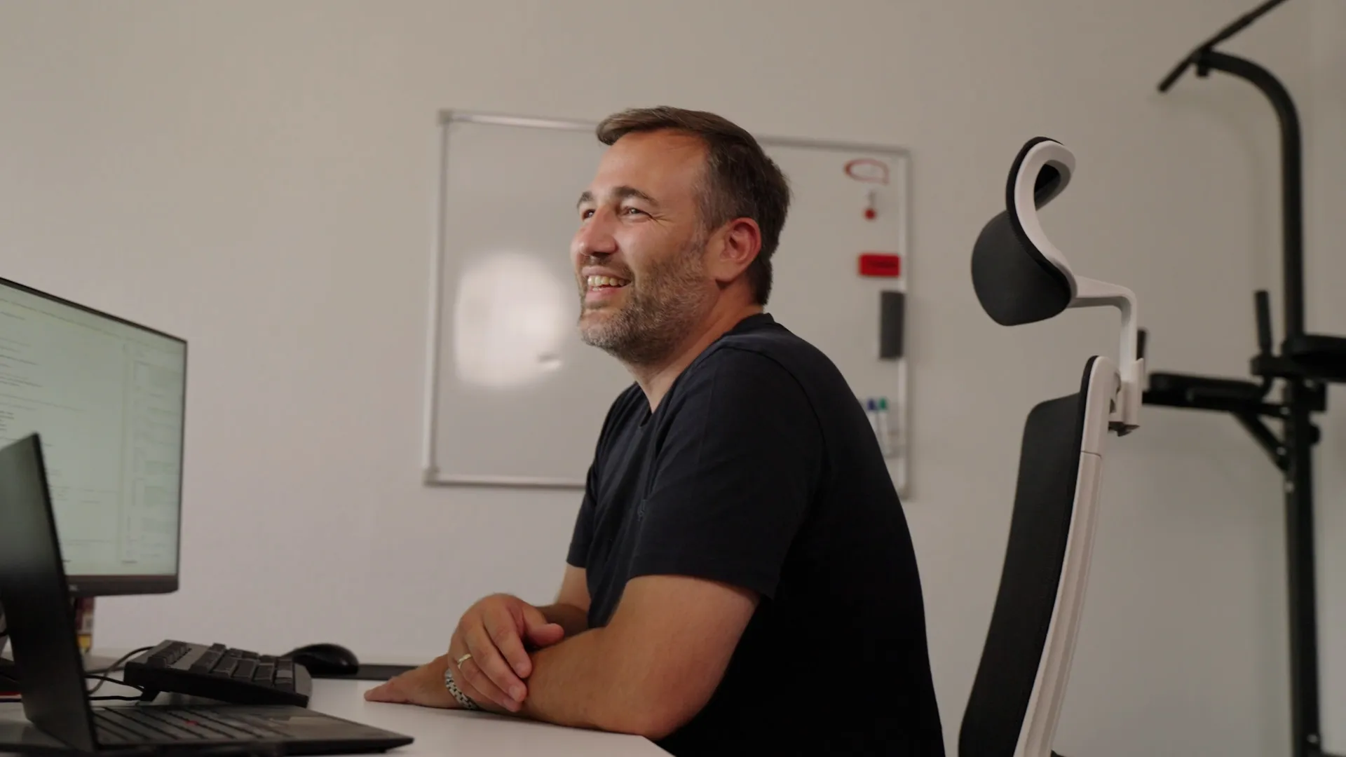 IDALABS-Gründer Tobias Gürtler sitzt auf einem ergonomischen Stuhl im IDALABS-Office. Er trägt ein schwarzes T-Shirt und schaut lächelnd über seinen Bildschirm. Im Hintergrund sind ein Whiteboard sowie eine Klimmzugstange zu erkennen.