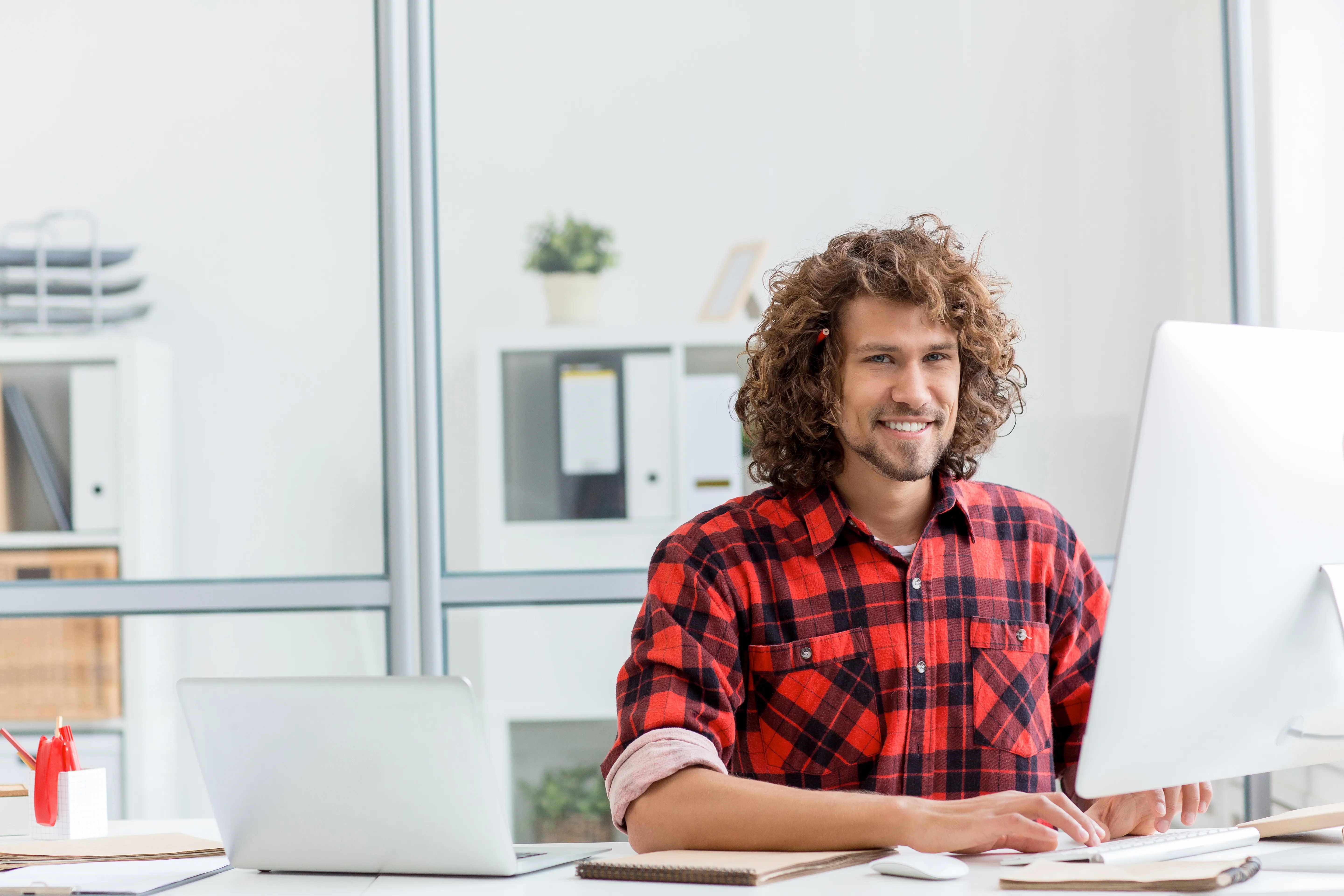 Ein junger Mann im karierten Hemd arbeitet an einem Computer. Er hat braune Locken und ihm klemmt ein roter Bleistift hinter seinem rechten Ohr.