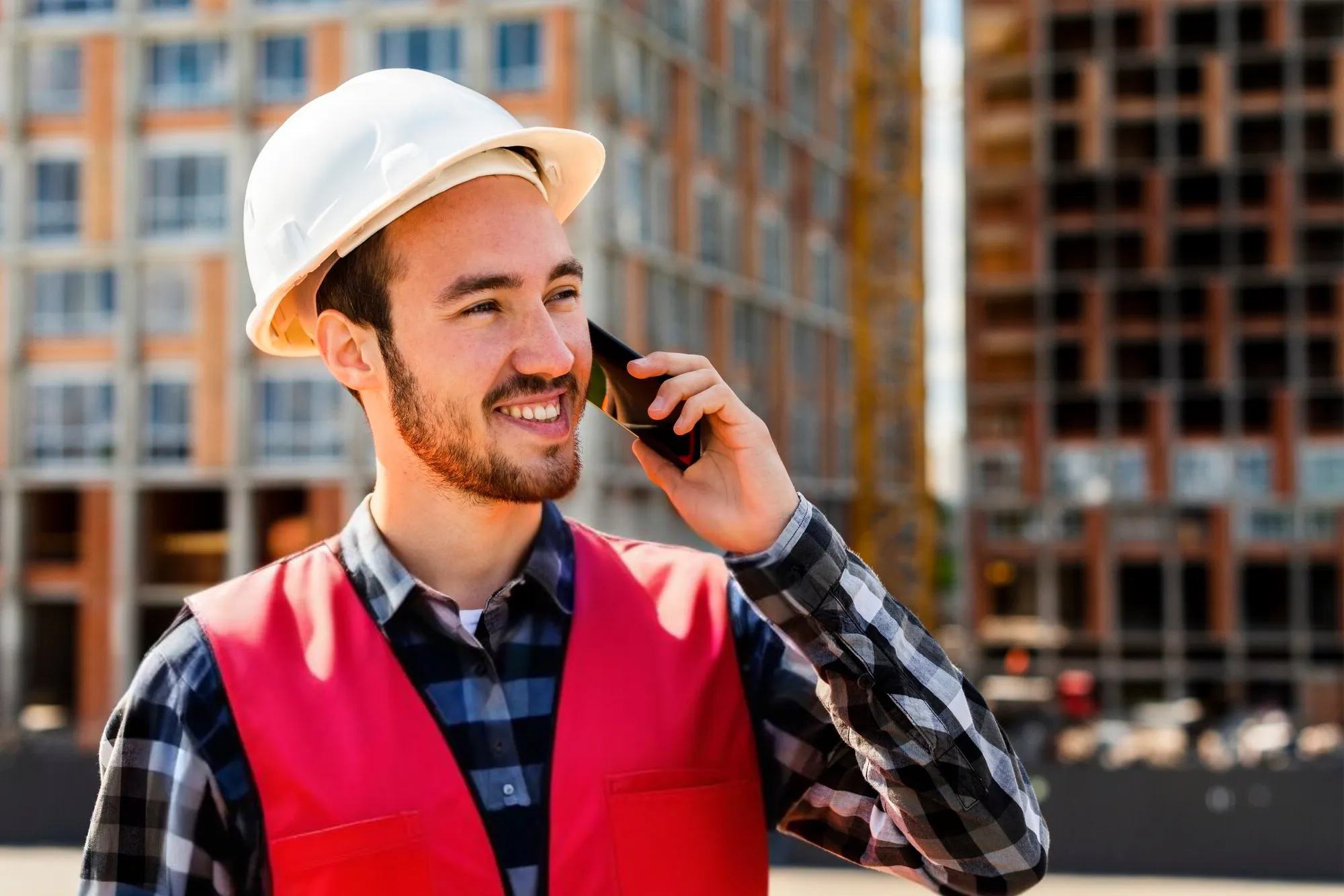Vor einem mehrstöckigen Gebäude mit Glasfront ist ein Mann zu sehen, der lächelnd ein Smartphone an sein Ohr hält. Er trägt ein kariertes Hemd sowie eine rote Warnweste und einen weißen Sicherheitshelm