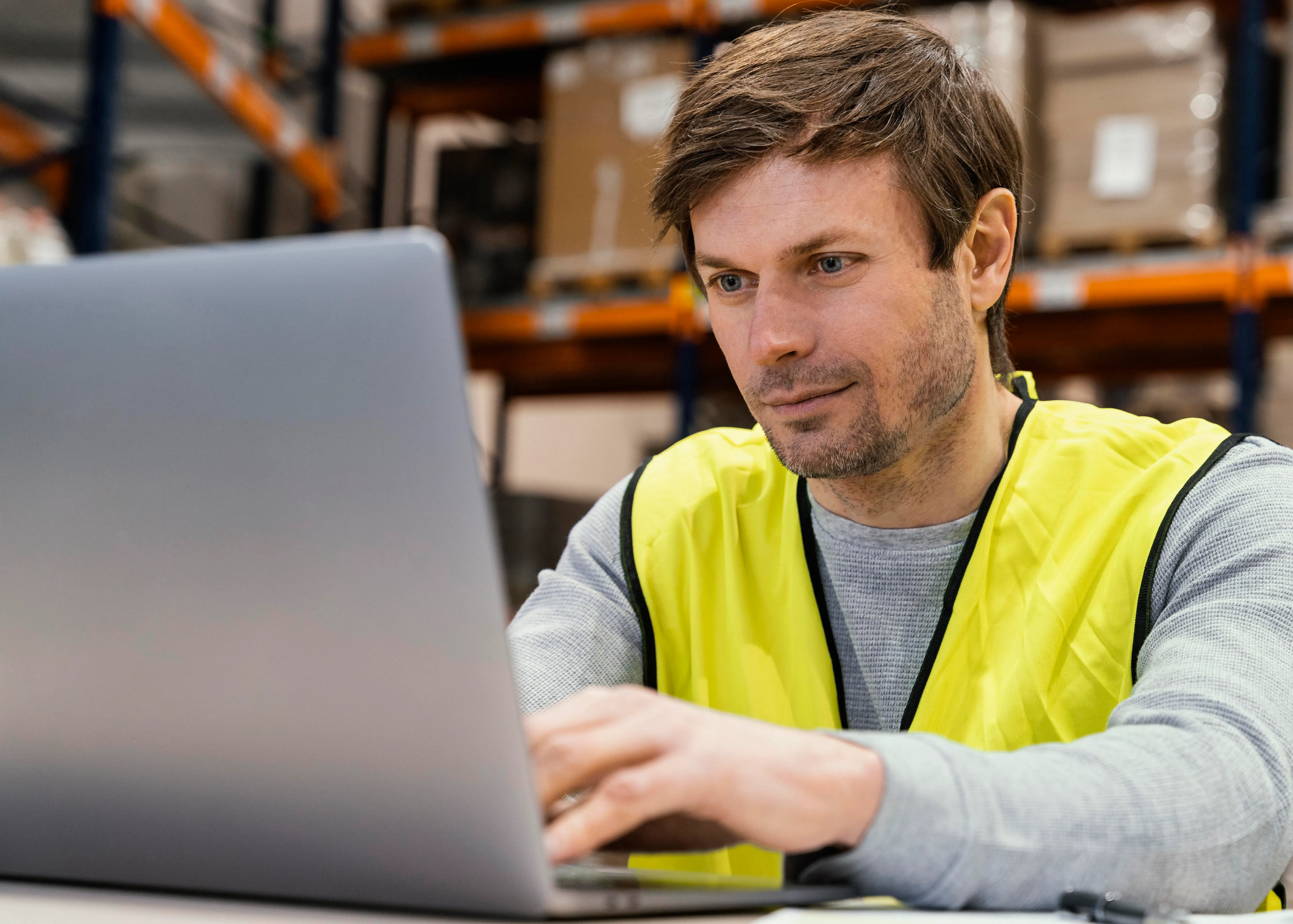 Ein Mann in gelber Warnweste arbeitet an einem Laptop. Im Hintergrund sind die hohen Regale eines Warenlagers zu sehen.