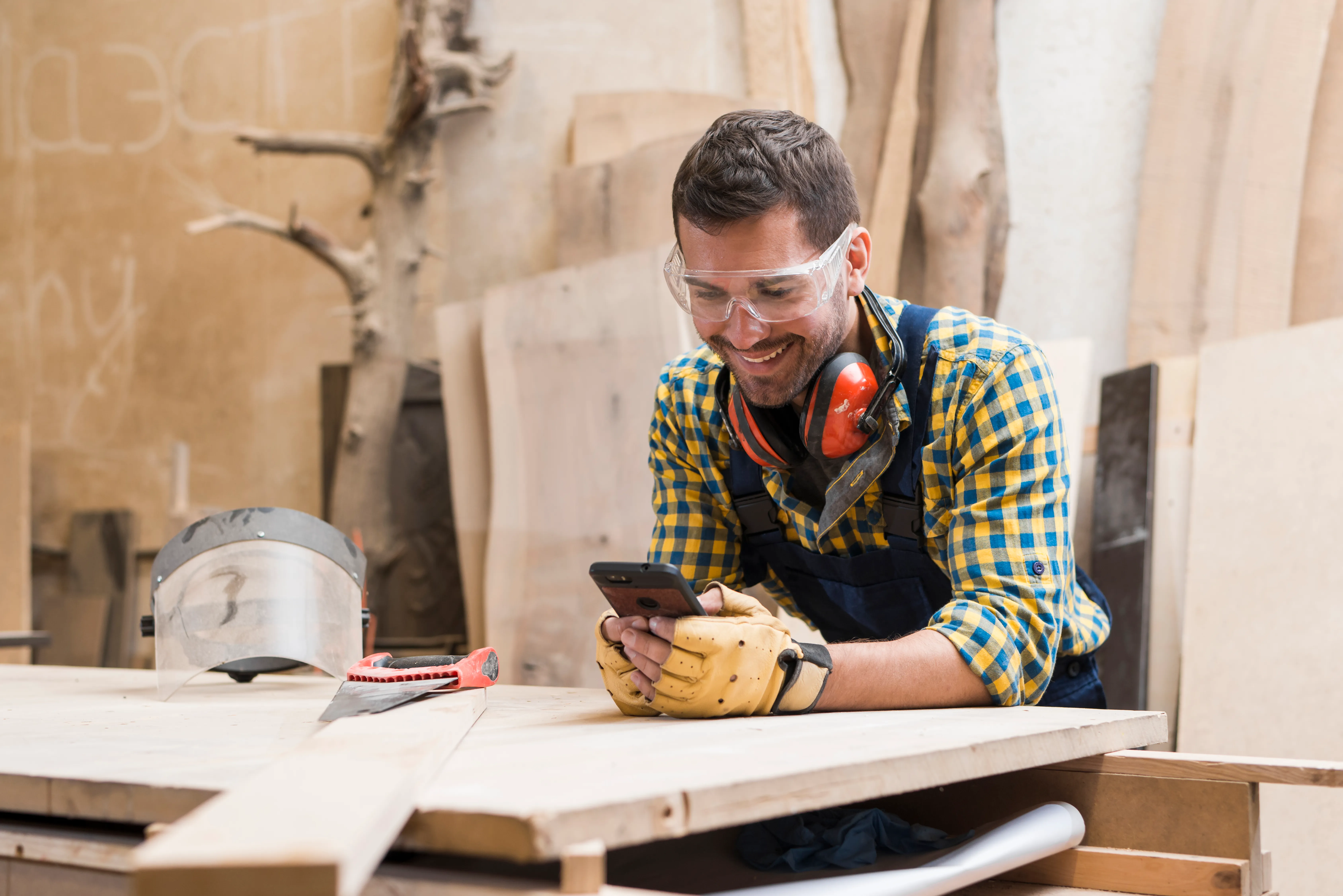 In einer Holzwerkstatt lehnt ein Mann an einem hölzernen Arbeitstisch. Er trägt ein kariertes Hemd sowie eine Schutzbrille auf dem Kopf und einen roten Gehörschutz um den Hals. Er schaut lächelnd auf das Display seines Smartphones.